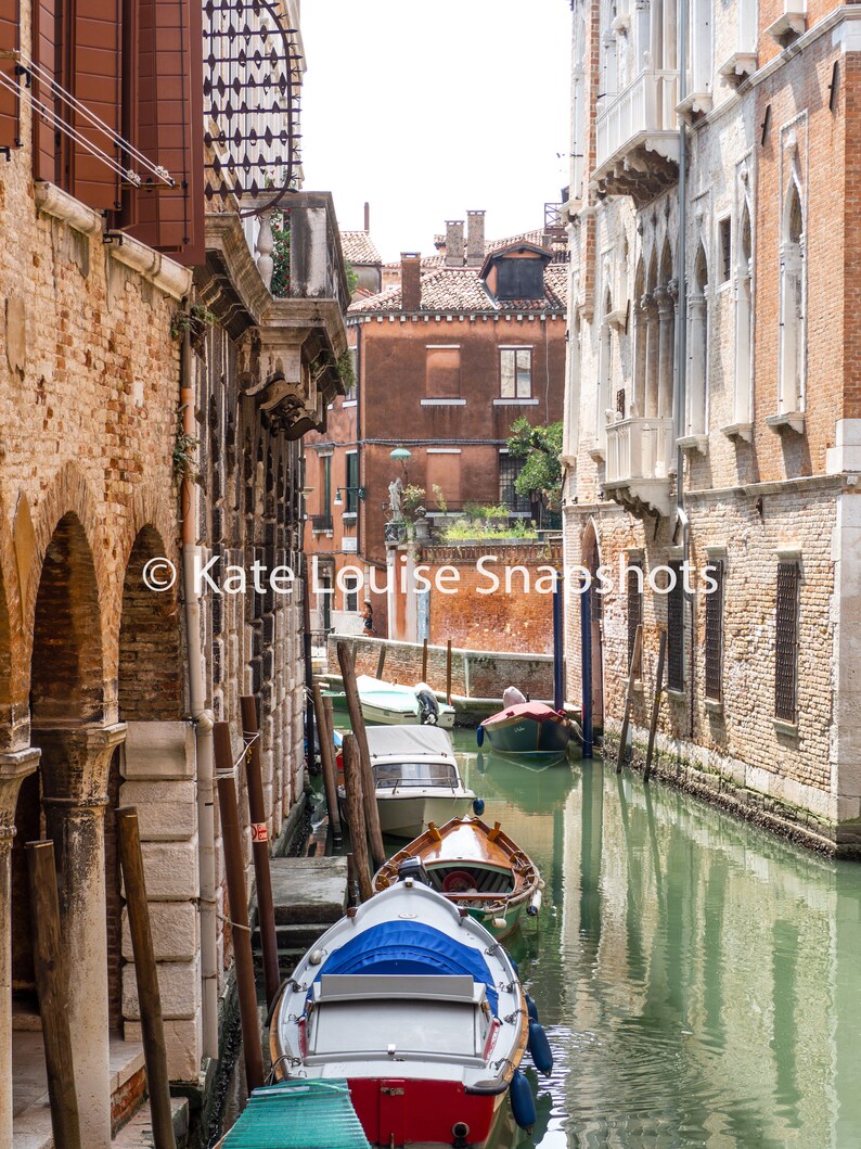Puede incluir: Una vista pintoresca de un canal veneciano con varios botes amarrados a lo largo del agua. Los edificios a ambos lados son de ladrillo y piedra, con ventanas arqueadas y balcones. El agua es verdosa y el cielo est&aacute; despejado.