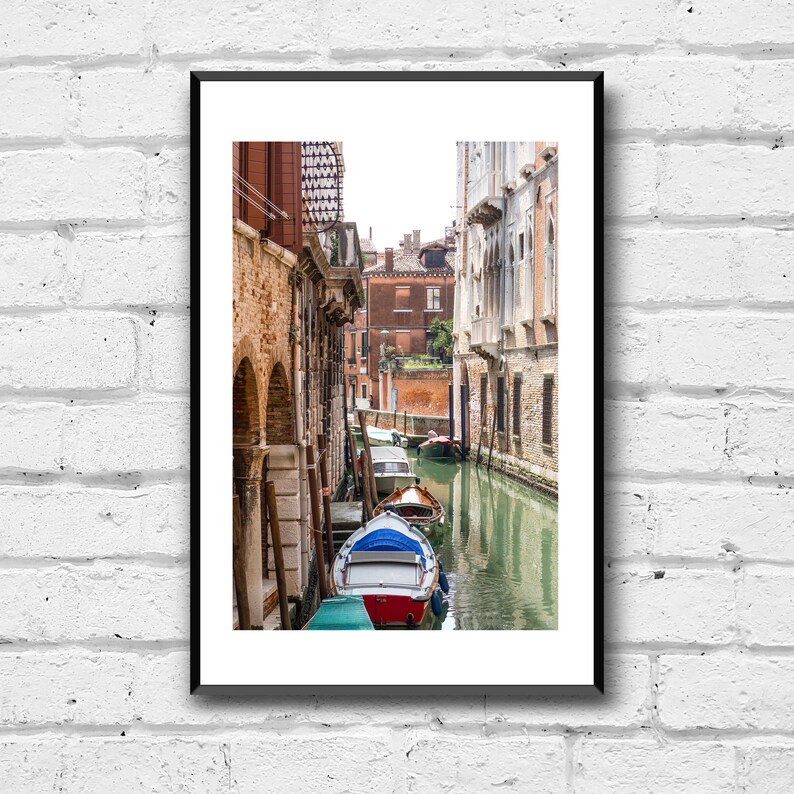 Puede incluir: Una impresi&oacute;n enmarcada de una escena del canal veneciano. La imagen presenta una v&iacute;a fluvial estrecha flanqueada por edificios de ladrillo envejecido. Varios botes est&aacute;n amarrados a lo largo del canal, con un bote rojo y blanco en primer plano. El agua refleja los edificios y el cielo.