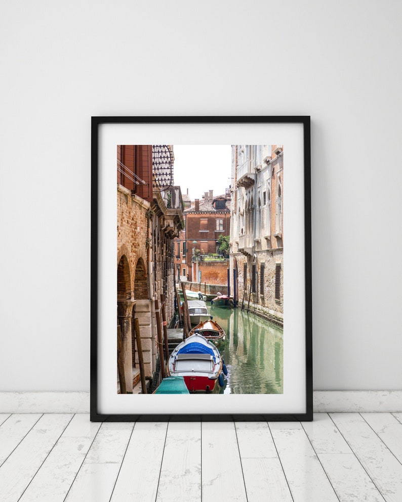 Puede incluir: Una impresi&oacute;n enmarcada de una escena del canal veneciano. La imagen presenta una v&iacute;a fluvial estrecha flanqueada por edificios de ladrillo envejecido. Un peque&ntilde;o bote con detalles rojos, blancos y azules est&aacute; en primer plano, con otros botes m&aacute;s abajo en el canal. El agua refleja los edificios y el cielo.