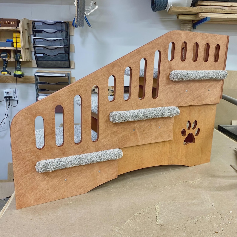 May include: A wooden pet staircase featuring three steps covered in gray speckled carpet. The side panel has vertical cutouts and a decorative paw print cutout. The structure is made of light-toned plywood with visible wood grain throughout.