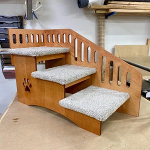 May include: A three-step pet staircase constructed from light brown wood with a smooth finish. Each step is covered in a textured, light gray carpet. The side panel features a decorative cutout of a paw print and a series of vertical oval slots.
