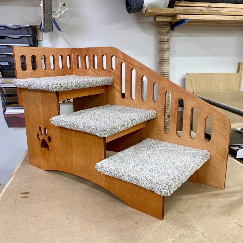 May include: A three-step pet staircase constructed from light brown wood with a smooth finish. Each step is covered in a textured, light gray carpet. The side panel features a decorative cutout of a paw print and a series of vertical oval slots.