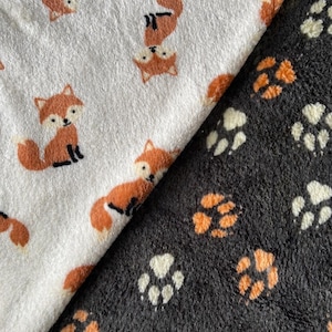 May include: Two plush fabrics, one white with orange foxes, the other black with orange and white paw prints. The fox fabric has repeating cartoon foxes. The paw print fabric has repeating paw prints.