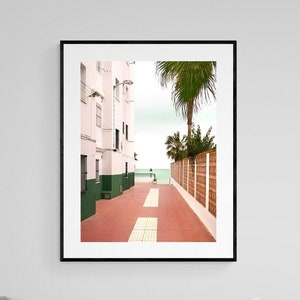 Puede incluir: Una fotografía de un camino estrecho de ladrillos rojos que conduce a una vista del océano. El camino está bordeado por edificios blancos a un lado y una valla de madera al otro. Se puede ver una palmera en el fondo.