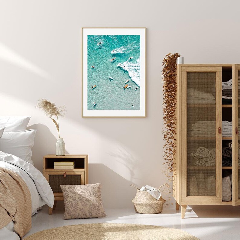 May include: An aerial photograph of a group of people surfing in turquoise water with white waves. The photo is framed in a light wood frame and hanging on a white wall.