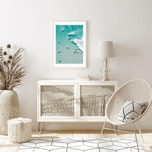 May include: A framed photograph of a beach scene with surfers in the water. The photo is in a white frame and is hanging on a white wall. The wall is also decorated with a white wicker chair and a white side table with a lamp.