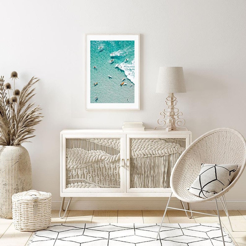 May include: A framed photograph of a beach scene with surfers in the water. The photo is in a white frame and is hanging on a white wall. The wall is also decorated with a white wicker chair and a white side table with a lamp.