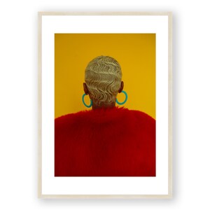 May include: A person with short, blonde, wavy hair wearing large, turquoise hoop earrings and a red fur coat against a yellow background.