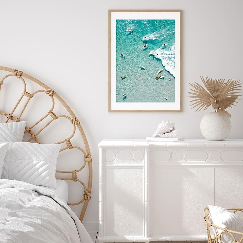 May include: An aerial photograph of a group of people surfing in turquoise water with white waves. The photo is framed in a light wood frame and hanging on a white wall.