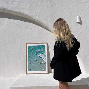 May include: A framed photograph of a beach scene with people surfing and swimming in turquoise water. The photo is hanging on a white wall.