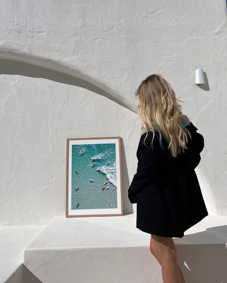May include: A framed photograph of a beach scene with people surfing and swimming in turquoise water. The photo is hanging on a white wall.