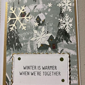 May include: A handmade winter greeting card with a gold border and a white rectangle with the text "WINTER IS WARMER WHEN WE'RE TOGETHER." The card features a snowy village scene and white snowflake cutouts.