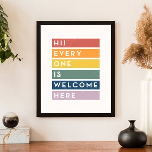 May include: A framed art print with the text "HI! EVERYONE IS WELCOME HERE" in colorful horizontal blocks. The print is in a black frame and is displayed on a wooden surface. A white vase with dried flowers and a black vase are also on the surface.