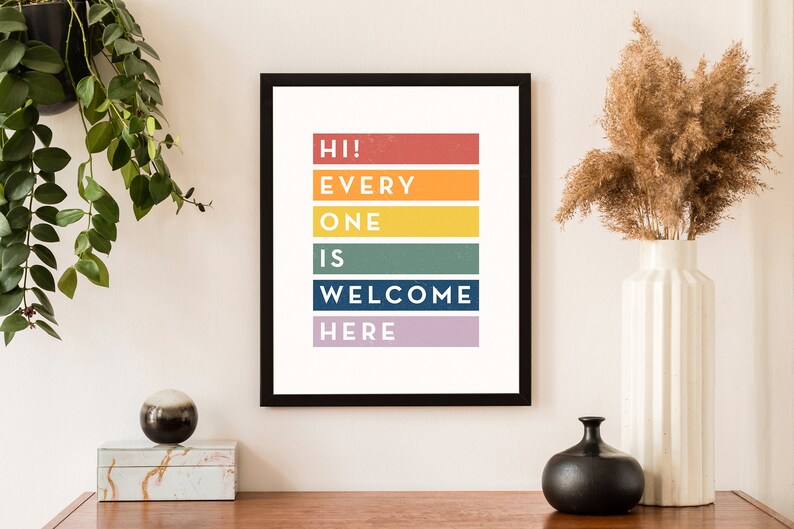 May include: A framed art print with the text "HI! EVERYONE IS WELCOME HERE" in colorful horizontal blocks. The print is in a black frame and is displayed on a wooden surface. A white vase with dried flowers and a black vase are also on the surface.