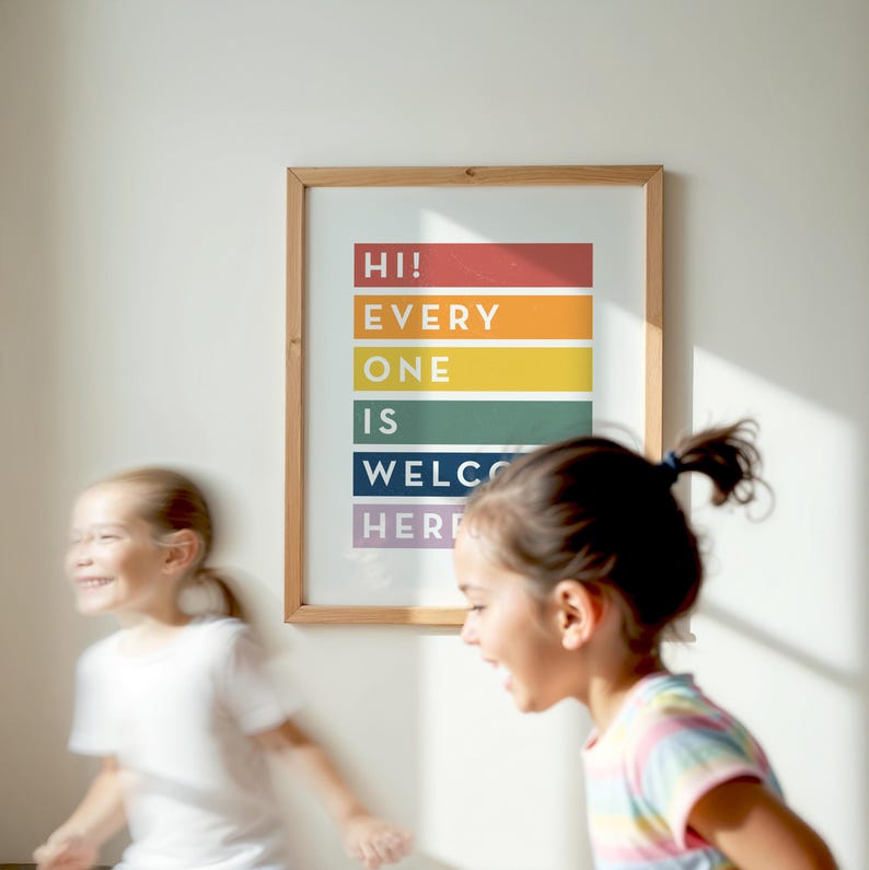 May include: A framed print with the words "HI! EVERYONE IS WELCOME HERE" in colorful vertical blocks. The frame is light brown. Two young girls are in the foreground, one wearing a white shirt and the other a striped shirt.