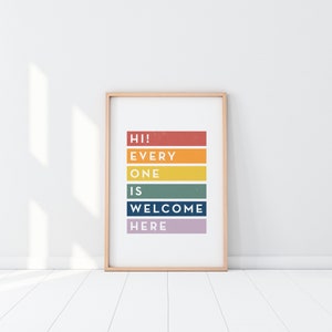 May include: A framed art print with the words "HI! EVERYONE IS WELCOME HERE" in a colorful, stacked design. The text is arranged on horizontal bars of red, orange, yellow, green, blue, and purple. The frame is a light wood color.