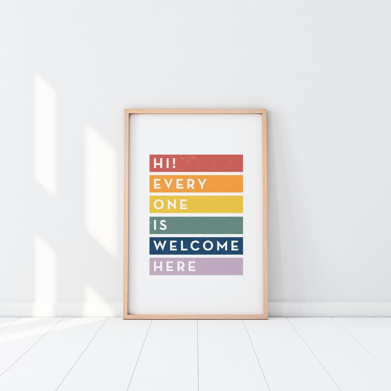 May include: A framed art print with the words "HI! EVERYONE IS WELCOME HERE" in a colorful, stacked design. The text is arranged on horizontal bars of red, orange, yellow, green, blue, and purple. The frame is a light wood color.