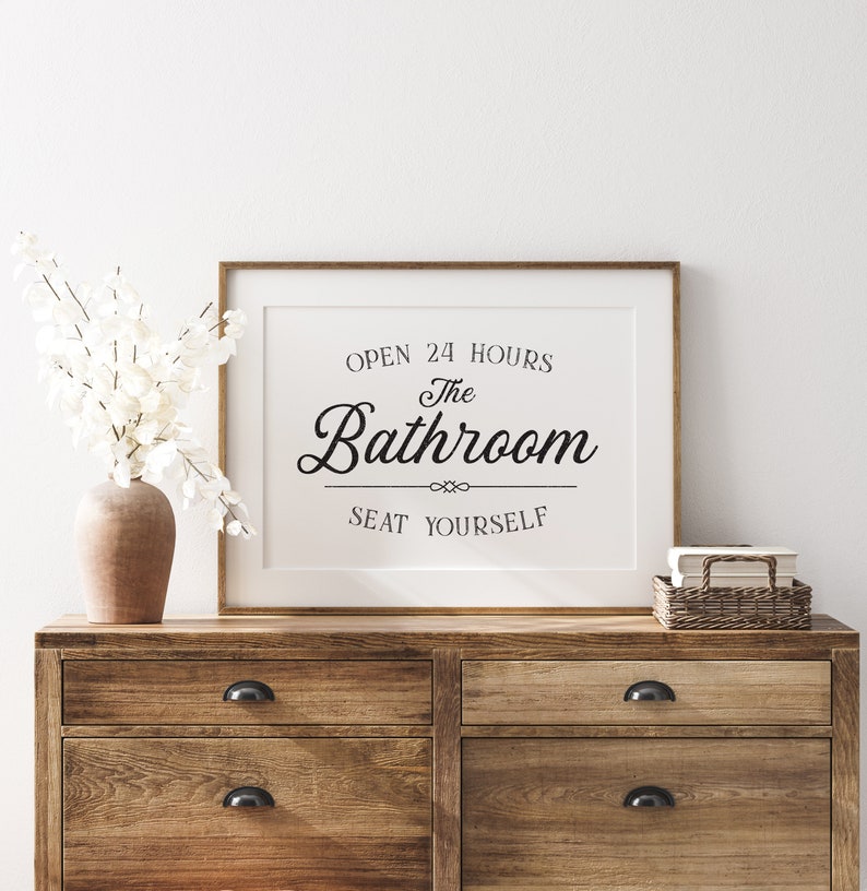May include: A framed print with the text "Open 24 Hours The Bathroom Seat Yourself" in a black cursive font. The print is on a white background and is sitting on a wooden dresser.
