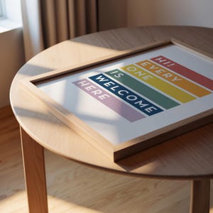 May include: A framed art print with the words "Hi! Every One Is Welcome Here" in a rainbow color scheme. The print is in a light wood frame, resting on a round wooden table. The artwork is a welcoming statement piece.
