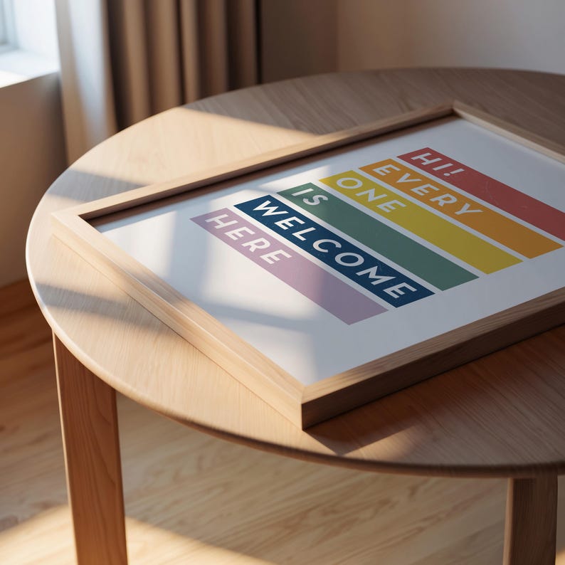 May include: A framed art print with the words "Hi! Every One Is Welcome Here" in a rainbow color scheme. The print is in a light wood frame, resting on a round wooden table. The artwork is a welcoming statement piece.