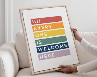 May include: A framed art print with the words "HI! EVERY ONE IS WELCOME HERE" in a rainbow color scheme. The print is held by a person on a white couch. The frame is a light wood color.