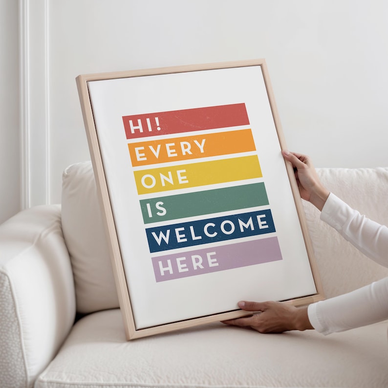 May include: A framed art print with the words "HI! EVERY ONE IS WELCOME HERE" in a rainbow color scheme. The print is held by a person on a white couch. The frame is a light wood color.