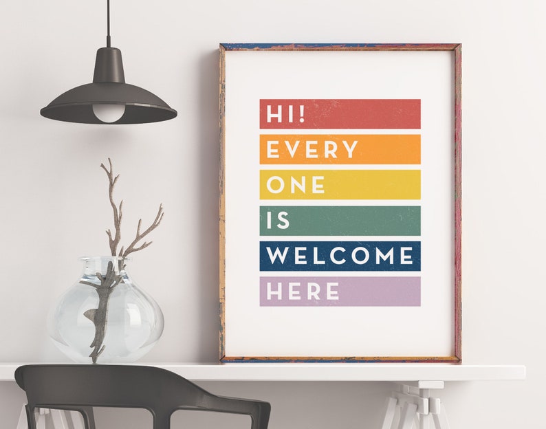 May include: A framed print with the words "HI! EVERYONE IS WELCOME HERE" in white text on colorful horizontal bars. The frame is a distressed blue and red. A glass vase with branches sits to the left.