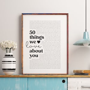 May include: A framed black and white print with the text "50 things we love about you" and a list of 50 reasons why someone is loved. The print is on a wooden shelf with a vase of white flowers and a lamp.