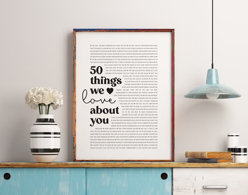 May include: A framed black and white print with the text "50 things we love about you" and a list of 50 reasons why someone is loved. The print is on a wooden shelf with a vase of white flowers and a lamp.
