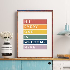 May include: A framed print with the words "HI! EVERYONE IS WELCOME HERE" in colorful horizontal blocks. The print is displayed on a blue cabinet with a vase of white flowers and a light blue pendant lamp.