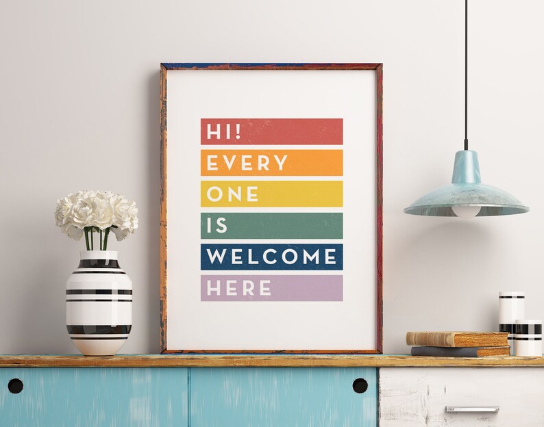 May include: A framed print with the words "HI! EVERYONE IS WELCOME HERE" in colorful horizontal blocks. The print is displayed on a blue cabinet with a vase of white flowers and a light blue pendant lamp.