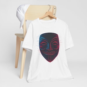 Op de afbeelding: Wit t-shirt met een gestileerd maskerontwerp in rood en blauw. De afbeelding lijkt op het Guy Fawkes-masker, een symbool van protest. Het t-shirt hangt aan een houten hanger.