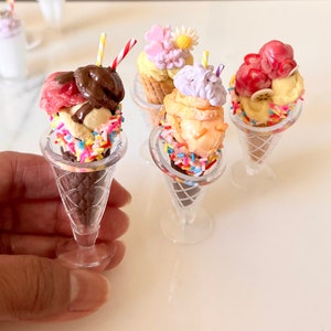 May include: Four miniature ice cream cones with different flavors and toppings. The cones are made of brown sugar cones and are filled with vanilla, strawberry, chocolate, and a pink and purple swirl ice cream. The cones are decorated with colorful sprinkles.
