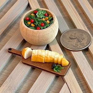 Puede incluir: Un bol de madera en miniatura lleno de una ensalada de lechuga verde, tomates cherry rojos y granos de maíz amarillos. Una tabla de cortar de madera con una pequeña hogaza de pan rebanado y aceitunas verdes está al lado del bol.