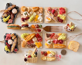 May include: A set of ten miniature charcuterie boards featuring a variety of cheeses, meats, and crackers. The boards are displayed on a white surface and include a fork and a small picnic basket.