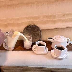 May include: A miniature white teapot with a lid, two white cups with saucers, and a clear pitcher pouring white liquid into a white glass. The items are arranged on a wooden surface.