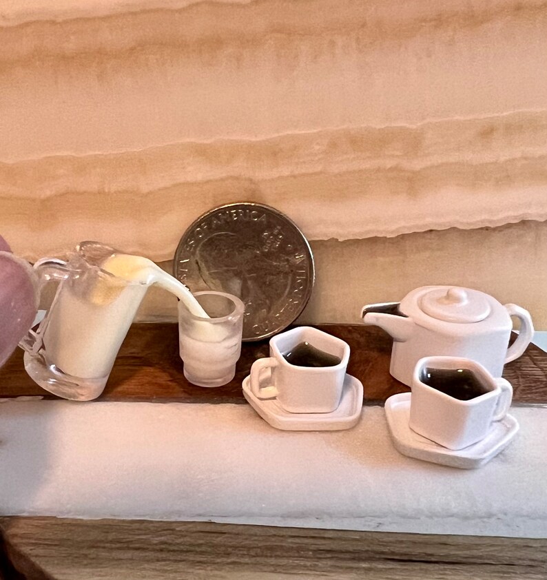 May include: A miniature white teapot with a lid, two white cups with saucers, and a clear pitcher pouring white liquid into a white glass. The items are arranged on a wooden surface.