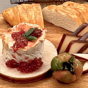 May include: A close-up of a wooden board with various food items. A round brie cheese topped with red jam and green leaves sits on a wooden plate. Sliced bread, cheese, and a green and orange fruit are also visible.