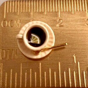 May include: A miniature teacup and saucer set with a gold spoon. The teacup is filled with dark tea and a lemon wedge. The set is on a gold-colored surface with a ruler.