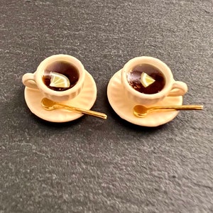 May include: Two white teacups with saucers and gold spoons. Each cup contains dark tea with a lemon wedge. The cups are on a dark gray surface.