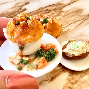 Pode incluir: Um conjunto em miniatura de três pratos com fundo branco. O primeiro prato contém um pastel de frango salgado com uma crosta dourada. O segundo prato tem um acompanhamento de feijão verde e cenoura. O terceiro tem uma batata assada com queijo e cebolinha.