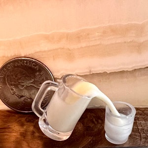 May include: A clear glass pitcher pouring a white liquid into a smaller clear glass. The pitcher has a handle and a lip. The liquid is pouring out of the pitcher and into the smaller glass. A silver coin is in the background.