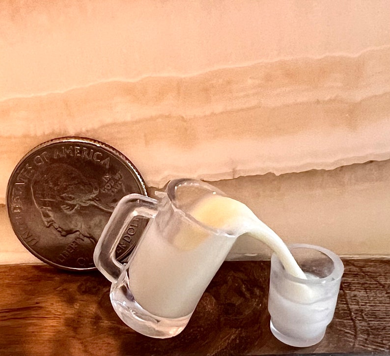 May include: A clear glass pitcher pouring a white liquid into a smaller clear glass. The pitcher has a handle and a lip. The liquid is pouring out of the pitcher and into the smaller glass. A silver coin is in the background.