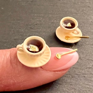 May include: Two miniature teacups and saucers with gold spoons. Each cup is filled with dark liquid and a lemon wedge. The cups are on a finger and a table.