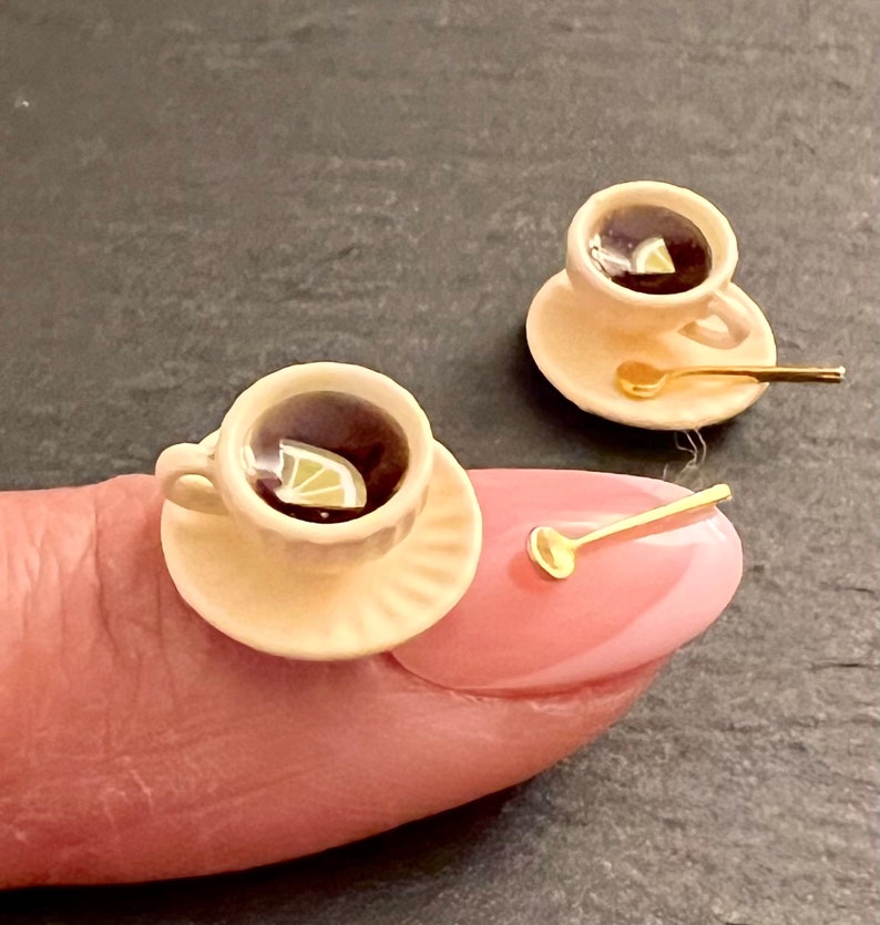 May include: Two miniature teacups and saucers with gold spoons. Each cup is filled with dark liquid and a lemon wedge. The cups are on a finger and a table.
