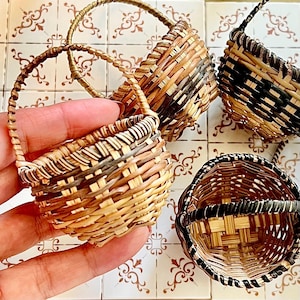 May include: A collection of miniature woven baskets in various shades of brown and black. Each basket has a small handle. The baskets are displayed on a patterned tile surface. The baskets are likely for decorative purposes.