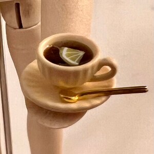 May include: A white teacup and saucer with a gold spoon. The teacup is filled with dark liquid and a slice of lemon.