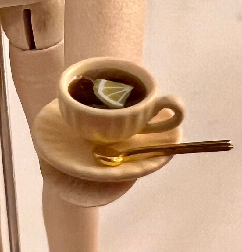 May include: A white teacup and saucer with a gold spoon. The teacup is filled with dark liquid and a slice of lemon.