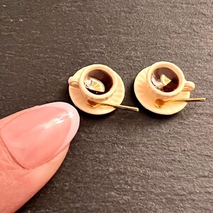 May include: Two miniature teacups with saucers and spoons. The cups are filled with brown liquid and have a lemon slice in each. The saucers are a light beige color and the spoons are gold.