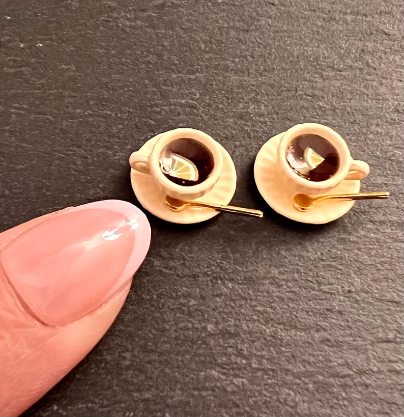 May include: Two miniature teacups with saucers and spoons. The cups are filled with brown liquid and have a lemon slice in each. The saucers are a light beige color and the spoons are gold.